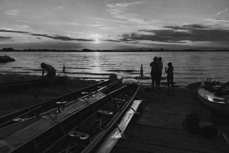 Ruderboote am Ufer bei Sonnenuntergang mit Menschen am Wasser.