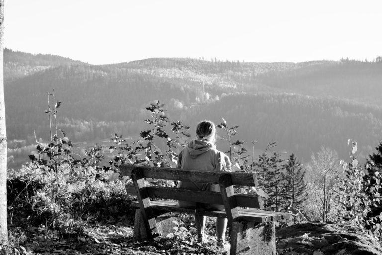 eine  Person sitzt auf einer Bank mit Blick auf bewaldete Hügel im Hintergrund.